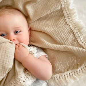 Op de afbeelding: Een beige muslin babydeken met witte franjes en de naam "Archie" geborduurd erop. Een baby ligt op de deken en houdt een hoek ervan in zijn mond.