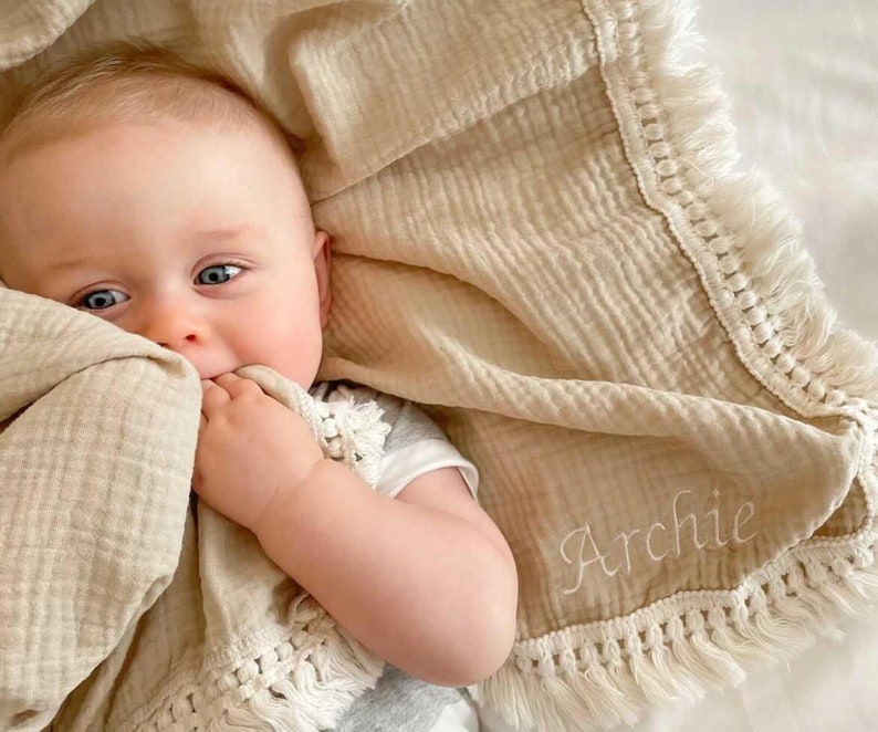 Op de afbeelding: Een beige muslin babydeken met witte franjes en de naam "Archie" geborduurd erop. Een baby ligt op de deken en houdt een hoek ervan in zijn mond.