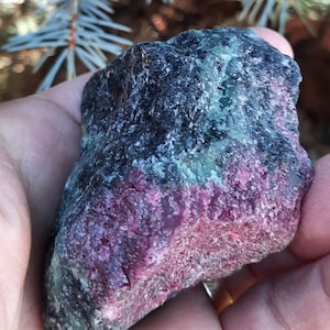 May include: A rough, dark grey, green, and pink gemstone with a rough, irregular shape.