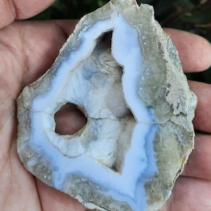 Puede incluir: Una geoda de ágata azul claro con un agujero en el centro. La geoda está rodeada por un exterior rugoso y gris.