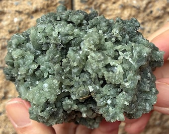 May include: A close-up of a cluster of green crystals. The crystals have a rough, textured surface and are clustered together in a dense formation. The color is a mix of light and dark green, with some areas appearing almost black. The overall shape is irregular and organic.