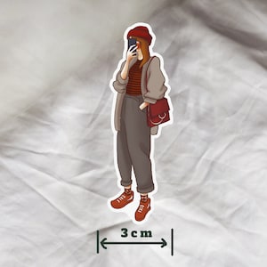 May include: A sticker of a person wearing a red hat, a striped sweater, and brown pants. The person is holding a phone and a brown bag with a circular handle. The sticker is 3 inches wide.