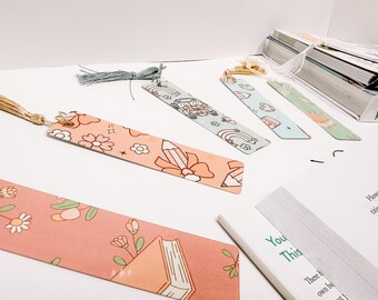 May include: A collection of colorful bookmarks with tassel accents. The bookmarks feature various designs, including floral patterns, pencils, and whimsical illustrations. They are arranged on a white surface near a stack of books.