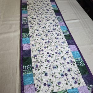 May include: Quilted table runner featuring a white floral pattern with purple and blue accents. The runner has a patchwork border in shades of purple, teal, and green, with a solid purple border. A decorative piece for a dining table.