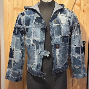Könnte beinhalten: Eine Patchwork-Jeansjacke mit grauer Kapuze. Die Jacke besteht aus verschiedenen Blautönen von Denim, die in einem Patchwork-Design zusammengenäht sind. Sie hat einen Knopfverschluss und Taschen. Der Markenname "UNIKALES" ist oben links sichtbar.