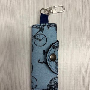 May include: A blue fabric keychain holder with a bicycle pattern. The holder has a snap closure and a silver-colored metal clasp. The fabric is light blue with black bicycle illustrations.