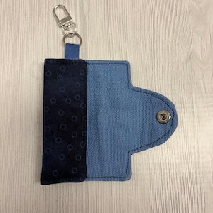 May include: A blue fabric key holder with a silver-colored clasp. The key holder is made of two different blue fabrics, one with a polka dot pattern. The key holder is secured with a snap closure. The brand name "UNIKALES" is visible in the top left corner.