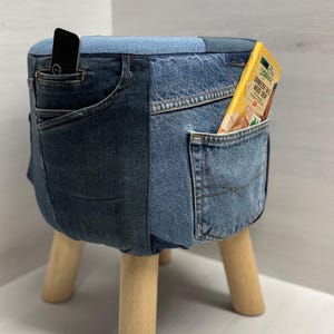 Puede incluir: Taburete cubierto de mezclilla con patas de madera clara. El taburete está hecho de jeans reciclados, con bolsillos en los laterales. Un teléfono inteligente y una caja de bocadillos son visibles en los bolsillos. Es un mueble único.