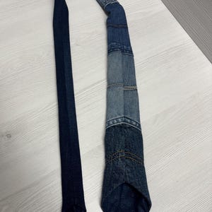 May include: A denim tie with a patchwork design. The tie features various shades of blue denim, with visible stitching and a patterned tip. The tie is laid out on a light-colored wooden surface.
