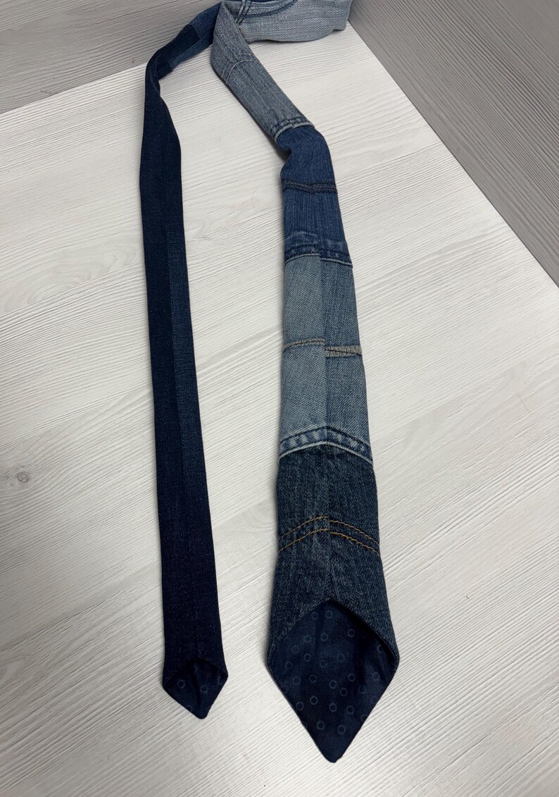 May include: A denim tie with a patchwork design. The tie features various shades of blue denim, with visible stitching and a patterned tip. The tie is laid out on a light-colored wooden surface.