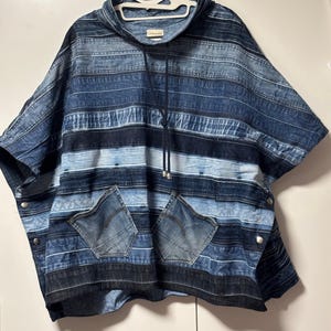 May include: A blue denim poncho with a hood and two front pockets. The poncho is constructed from various shades of blue denim, forming a striped design. It features silver snap fastenings on the sides and a drawstring at the neckline. The brand name "UNIKALES" is visible.