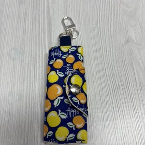 May include: A navy blue key holder with a pattern of yellow and orange apples and green leaves. The key holder has a silver snap closure and a silver key ring. The word "Apple" is printed on the key holder.