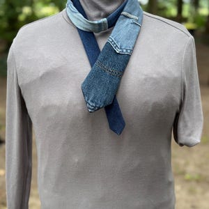May include: A denim scarf with a patchwork design, featuring various shades of blue. The scarf is draped over a gray turtleneck sweater, creating a layered look. The background is blurred, suggesting an outdoor setting.