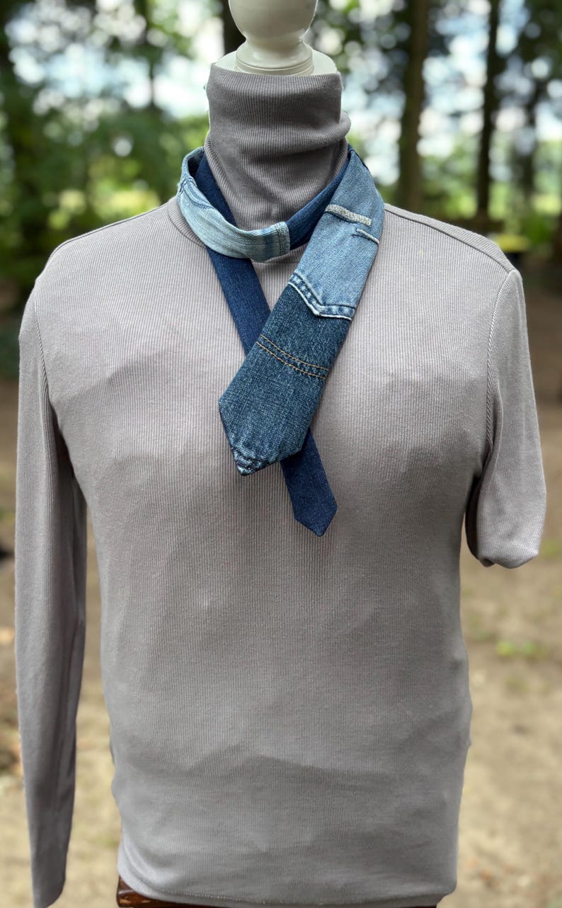 May include: A denim scarf with a patchwork design, featuring various shades of blue. The scarf is draped over a gray turtleneck sweater, creating a layered look. The background is blurred, suggesting an outdoor setting.