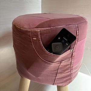 May include: A round stool upholstered in dusty pink denim fabric with visible seams and a functional pocket on the side. A black remote control is tucked into the pocket. The stool is supported by three light-coloured wooden legs.