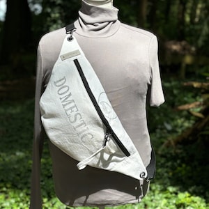 Puede incluir: Riñonera blanca de lona con cremallera y correa negras. La bolsa tiene la palabra "DOMESTIC" impresa en ella en una fuente gris.