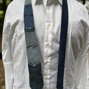 May include: A denim tie with a patchwork design, featuring various shades of blue denim. The tie is paired with a white dress shirt. The tie's design includes pocket details and contrasting stitching, adding a unique, handcrafted touch to the accessory.