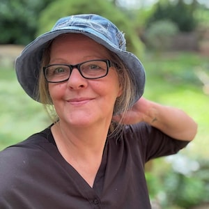 Op de afbeelding: Een lichtblauwe denim vissershoed met een gerafelde rand. De hoed wordt gedragen door een persoon die een bril met zwart montuur en een zwart overhemd draagt. De achtergrond is een vage groene buitenscène.