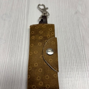 May include: A brown fabric keychain holder with a repeating circular pattern. The holder has a flap secured by a silver snap and a metal clasp for attaching keys. The brand name "UNIKALES" is visible in the top left corner.