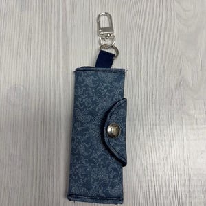 May include: A blue floral patterned key holder with a silver-colored clasp and a navy blue fabric loop. The key holder is closed with a silver button. The brand name "UNIKALES" is printed at the top left.