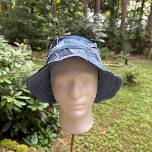 Op de afbeelding: Een denim patchwork bucket hat met een brede rand. De hoed is gemaakt van verschillende tinten blauwe denim, waardoor een getextureerd, meerkleurig uiterlijk ontstaat. De hoed wordt getoond op een mannequinhoofd tegen een achtergrond van groen gebladerte.