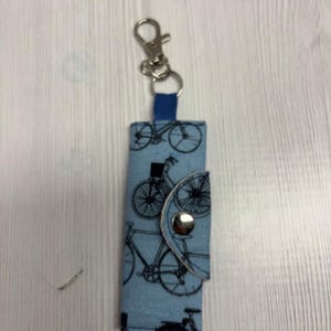 May include: A blue fabric keychain holder with a bicycle pattern. The holder has a silver metal clasp and a snap closure. The word "UNIKALES" is printed in the upper left corner.