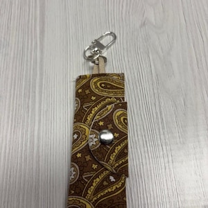 May include: A brown paisley print keychain holder with a silver clasp and snap closure. The fabric design features yellow and white accents. The brand name "UNIKALES" is visible in the upper left corner.