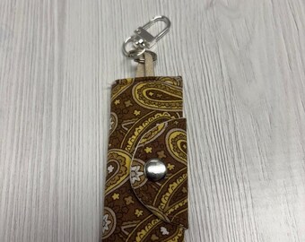 May include: A brown paisley print keychain holder with a silver clasp and snap closure. The fabric design features yellow and white accents. The brand name "UNIKALES" is visible in the upper left corner.