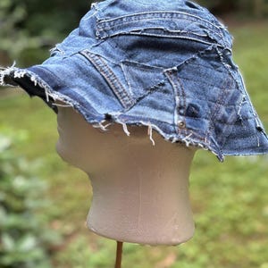 Può includere: Un cappello a secchiello in denim blu con bordo sfilacciato. Il cappello è realizzato con pezzi di denim, con cuciture a vista e un design patchwork. Il cappello è esposto su una testa di manichino.