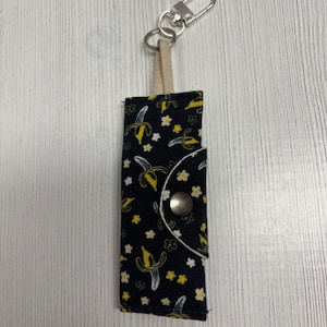 May include: A black fabric keychain holder with a banana pattern. The holder has a silver-colored metal clasp and a beige leather strap. The banana pattern includes yellow bananas and small white flowers.