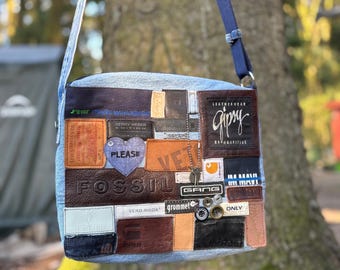May include: A light blue denim crossbody bag featuring a patchwork design of various leather and fabric brand labels, including Fossil, Gipsy, and Vero Moda. The bag includes a blue shoulder strap, a purple heart patch, and several metal buttons.