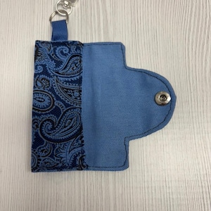 May include: Blue key holder with a paisley pattern on one side and a snap closure. The key holder has a silver-colored metal clasp and a small fabric loop for attachment. The word "UNIKALES" is printed at the top.