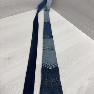 May include: A denim tie with a patchwork design. The tie features various shades of blue denim, with a darker blue section at the bottom and a lighter blue section at the top. The tie is lying on a light-colored surface.