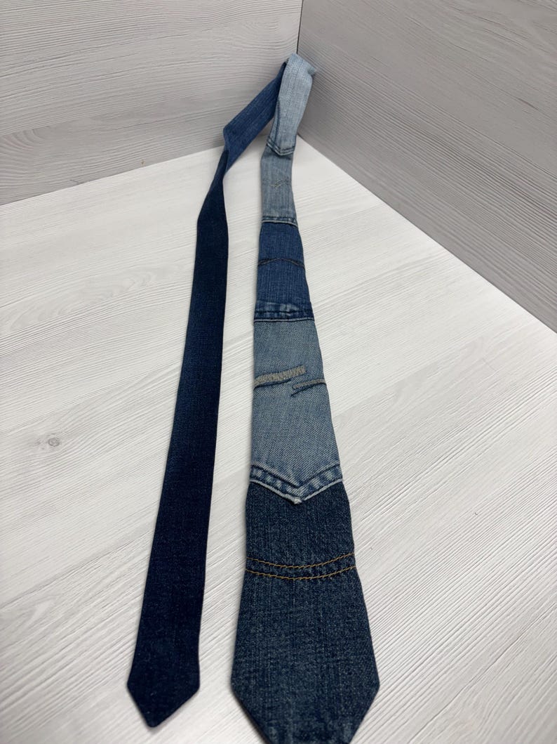 May include: A denim tie with a patchwork design. The tie features various shades of blue denim, with a darker blue section at the bottom and a lighter blue section at the top. The tie is lying on a light-colored surface.