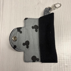 May include: A gray and black key holder with a snap closure and a silver-colored clasp. The gray fabric features a pattern of black Mickey Mouse silhouettes. The key holder is open, revealing a black interior.