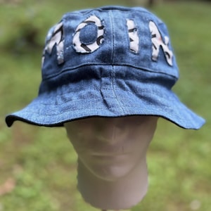 Könnte beinhalten: Ein blauer Denim-Eimerhut mit dem Wort "MOIA", das auf der Vorderseite aus weißem Stoff mit einem schwarz-weißen Muster appliziert ist. Der Hut wird auf einem weißen Schaufensterkopf vor einem verschwommenen grünen Hintergrund präsentiert.