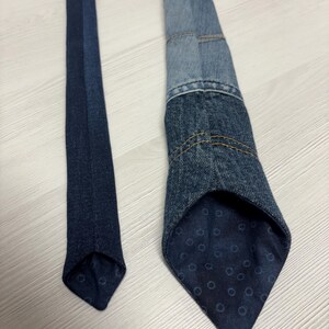 May include: A denim tie with a patchwork design, featuring various shades of blue denim. The tie's interior lining has a dark blue pattern with small circles. The tie is a classic accessory.
