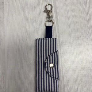 May include: A blue and white striped fabric keychain holder with a silver-colored clasp and a navy blue fabric loop. The holder has a flap with a silver snap closure. The brand name "UNIKALES" is visible in the top left corner.