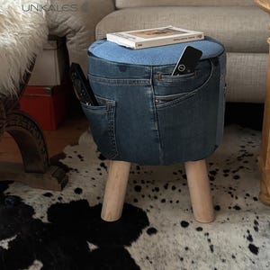 Puede incluir: Taburete cubierto de mezclilla con tela azul claro en la parte superior. El taburete tiene tres patas de madera y bolsillos de jeans en los lados. Un libro y un mando a distancia descansan encima. El taburete es un mueble único.