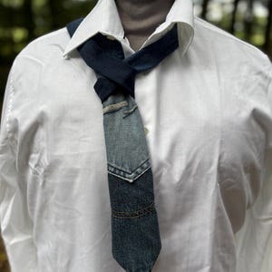 May include: A denim tie with a patchwork design, featuring various shades of blue. The tie is knotted and worn over a white collared shirt and a gray turtleneck. The denim appears to be from recycled jeans.