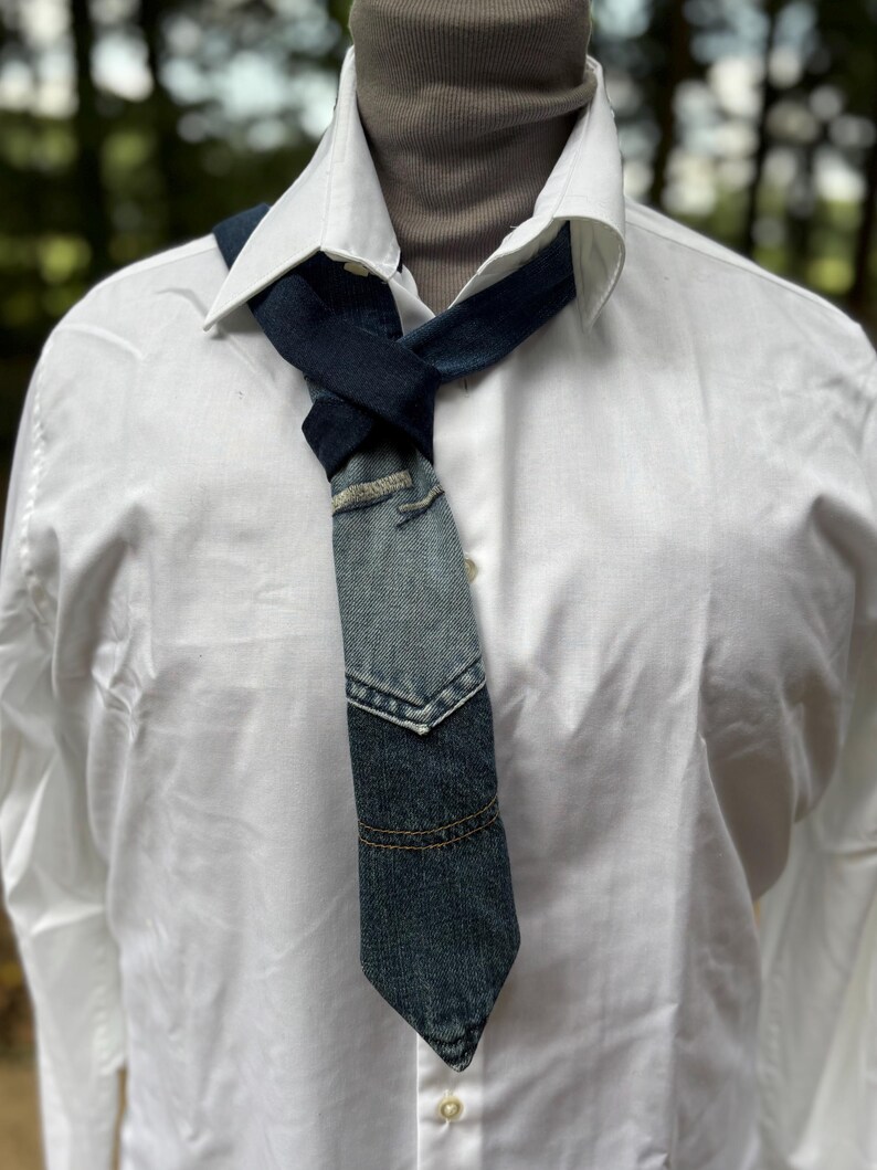 May include: A denim tie with a patchwork design, featuring various shades of blue. The tie is knotted and worn over a white collared shirt and a gray turtleneck. The denim appears to be from recycled jeans.