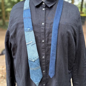 May include: A denim tie with a patchwork design, featuring various shades of blue. The tie is worn over a black button-down shirt. The patchwork includes a pocket detail, adding a unique touch to the accessory. The tie's design suggests a handcrafted or upcycled aesthetic.