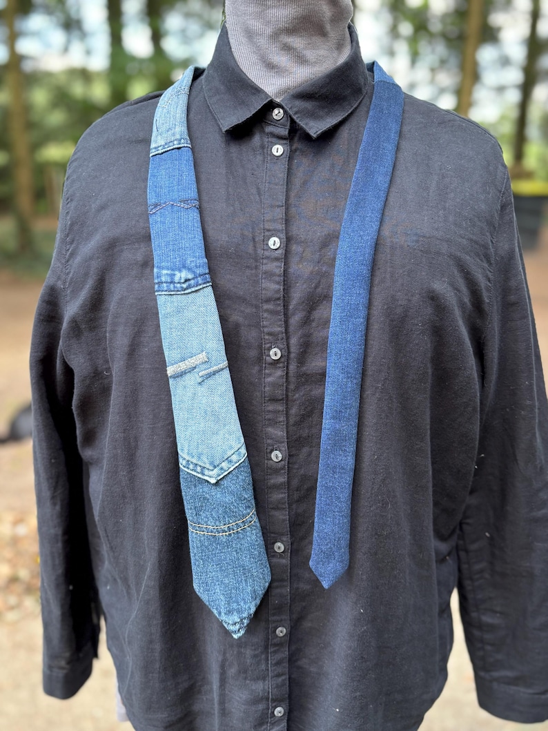 May include: A denim tie with a patchwork design, featuring various shades of blue. The tie is worn over a black button-down shirt. The patchwork includes a pocket detail, adding a unique touch to the accessory. The tie's design suggests a handcrafted or upcycled aesthetic.