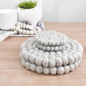 Op de afbeelding: Drie grijze vilt onderzetters gestapeld op elkaar. De onderzetters zijn gemaakt van kleine, ronde viltballen. De onderzetters staan op een houten tafel.