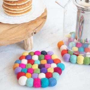 Può includere: Due sottobicchieri rotondi in feltro colorati, realizzati con piccole sfere di filato in vari colori, tra cui rosso, arancione, giallo, verde, blu, viola e bianco. I sottobicchieri sono su una superficie bianca.