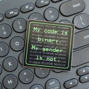 Może przedstawiać: Czarno-biała naklejka z zielonym tekstem, który brzmi "My code is binary. My gender is not." Naklejka jest umieszczona na szarej klawiaturze.