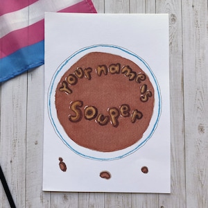 Puede incluir: Impresión artística en acuarela que presenta un círculo marrón con las palabras "your name is Souper" en letras marrones. La obra de arte tiene un borde azul y está sobre papel blanco. Una bandera del orgullo transgénero es visible.
