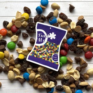 Puede incluir: Una bolsa morada y blanca con el texto "Trail Mix" y "Now with M&Ms" en ella. La bolsa está llena de caramelos y nueces de colores. La bolsa está rodeada de una variedad de nueces, caramelos y chispas de chocolate.