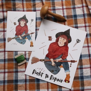 May include: Two art prints featuring a witch riding a broom, with the text "Right to Repair." The witch wears a black hat, red sweater, and blue jeans. A wooden spool of green thread and a wooden tool are also present.