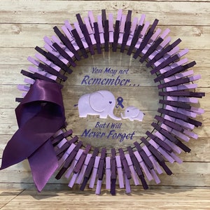 May include: Purple and lavender painted wooden clothespins arranged in a circle to form a wreath. A purple ribbon is tied to the wreath. The center of the wreath features a white background with the text "You May not Remember... But I Will Never Forget" and two purple elephants with a purple ribbon.